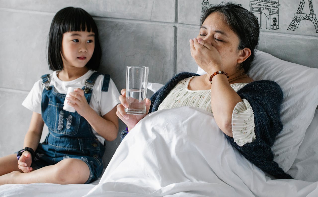Cute Asian little girl sitting on soft bed and looking at sick grandmother drinking medicine on sunny day