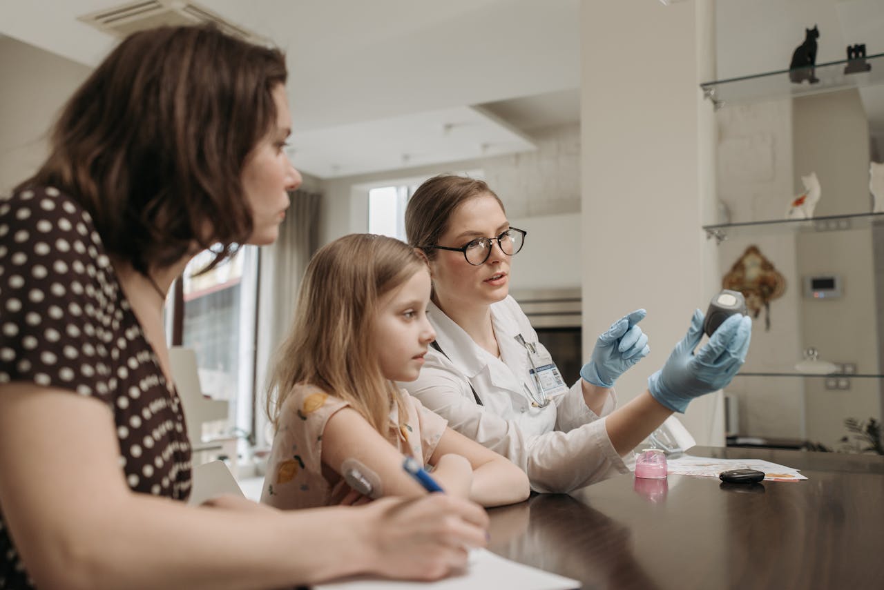 A doctor explains glucometer usage to a mother and daughter during a home visit.