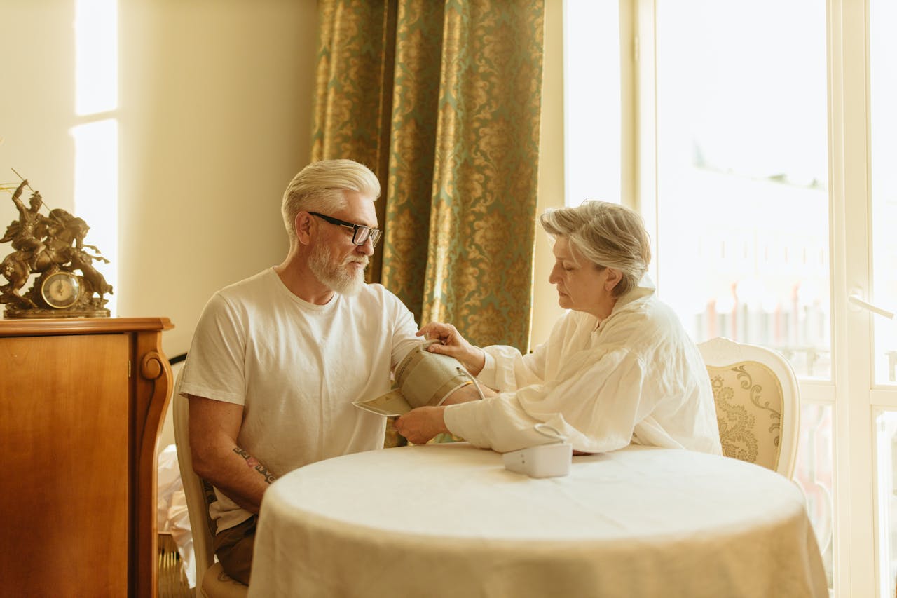 An elderly couple checks blood pressure using a digital monitor at their home table.